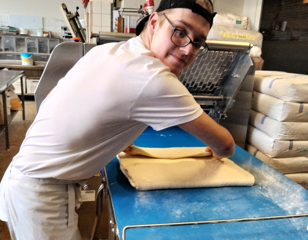 Ein junger Mann in weißer Bäcker-Arbeitskleidung hält einen Teigroller in der Hand und rollt einen Teig aus