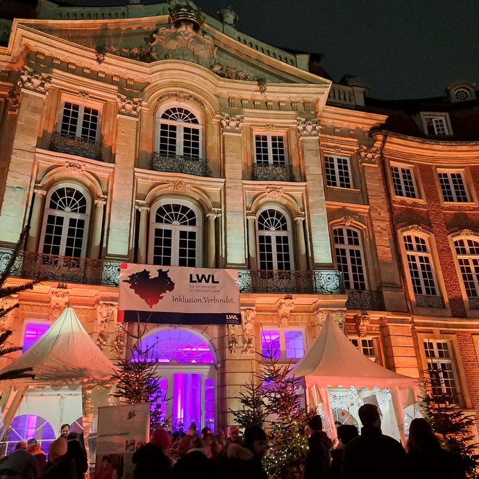 Der historische Erbdrostenhof in Münster mit Lichtern beleuchtet. Davor stehen Tannenbäume und Weihnachtsmarktstände. (vergrößerte Bildansicht wird geöffnet)
