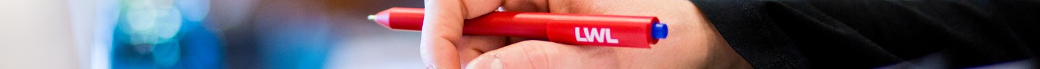 Verschwommener Hintergrund. Im Fokus eine Hand die einen roten LWL-Kugelschreiber hält.