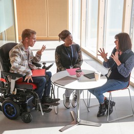 Zwei Frauen und ein Mann sitzen um einen runden Tisch herum. Der Mann sitzt im Rollstuhl. Alle drei unterhalten sich angeregt.