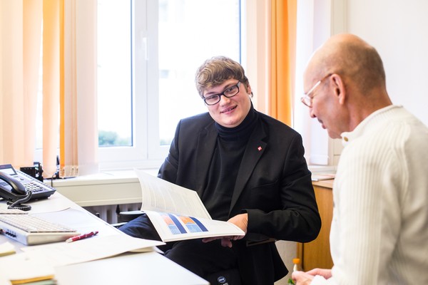 Ein Mann sitzt am Schreibtisch und spricht mit einem anderen Mann, der im Rollstuhl sitzt.