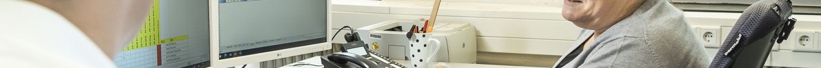 Eine Frau im Rollstuhl arbeitet an einem Schreibtisch in einem Büro. Sie schaut freundlich zu einem Kollegen, der neben ihr sitzt.